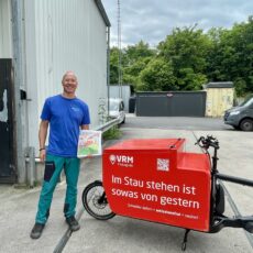 Thomas Schmitz von der Firma VRM City Logistik steht neben rotem VRM-Lastenrad mit Aufschrift „Im Stau stehen ist sowas von gestern“ und hält einen Sensor in der Hand, auf einem Betriebshof in Wiesbaden.